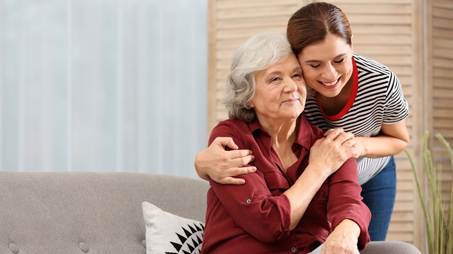 Frau nimmt eine auf dem Sofa sitzende Seniorin in den Arm Frau nimmt eine auf dem Sofa sitzende Seniorin in den Arm